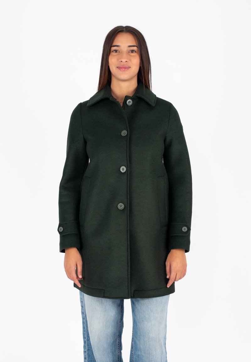 Cappotto di lana verde scuro con colletto strutturato, chiusura a bottoni e tasche laterali; le maniche hanno polsini con bottoni; indossato sopra jeans azzurri.