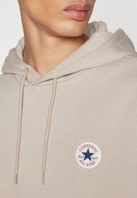 Beige hettegenser laget av myk stoff, med en sirkulær "Converse All Star" logo med en blå stjerne og røde detaljer.