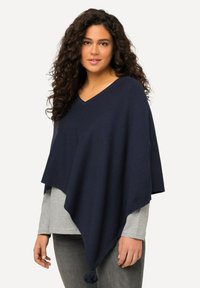Navy sininen neuleponcho v-pääntiellä, jossa on tupsu korostus helmassa, ja se on kerroksittain vaaleanharmaan pitkähihainen paidan päällä. Pehmeä rakenne.