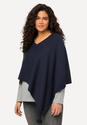Poncho en tricot bleu marine avec un col en V, accent de glands à l'ourlet et superposé sur une chemise à manches longues gris clair. Texture douce.
