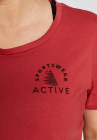 Camiseta de algodón roja con cuello redondo, que presenta texto bordado en negro que dice "SPORTSWEAR ACTIVE" y un patrón sobre el texto.