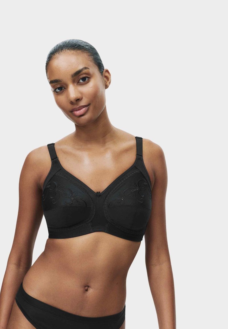 Donna con capelli raccolti che indossa un reggiseno nero ricamato e biancheria intima abbinata, in piedi su uno sfondo chiaro e uniforme.