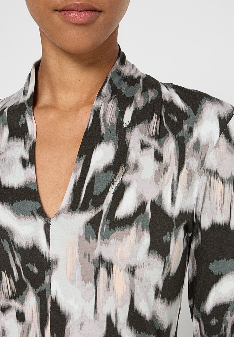 Gemusterte Bluse mit V-Ausschnitt. Präsentiert abstrakte graue, weiße und pfirsichfarbene Formen auf einem dunklen Hintergrund. Das Material scheint weich und strukturiert zu sein.