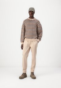 Beige strikket sweater med mønsterdesign, lyse beige bukser og lysebrune snørebåndsstøvler. Modellen bærer en lys grå kasket.