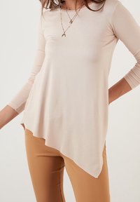 Top lungo beige chiaro a maniche lunghe con orlo asimmetrico, realizzato in un tessuto morbido; indossato su pantaloni aderenti color marrone e stratificato con collane.