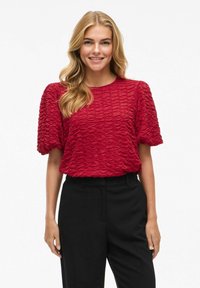 Blouse rouge texturée avec des manches courtes bouffantes et un col rond, assortie à un pantalon noir. Le tissu présente un motif ondulé.