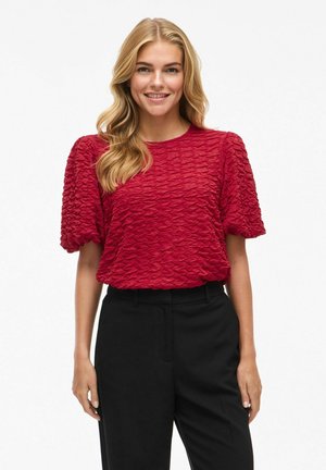 Blouse rouge texturée avec des manches courtes bouffantes et un col rond, assortie à un pantalon noir. Le tissu présente un motif ondulé.