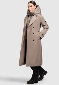 Beige trenchcoat met een afneembare gewatteerde capuchon, dubbele knopen aan de voorkant en zwarte knopen, gecombineerd met zwarte broek en chunky laarzen.