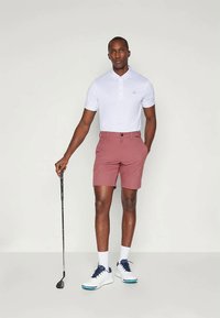 White polo shirt, pink shorts, and white golf shoes with blue accents. Standing with a golf club, neutral background, focused pose.