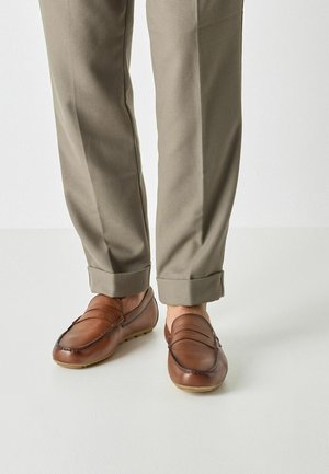 Mocassini in pelle marrone con dettagli cuciti e design senza lacci. Indossati con pantaloni grigio chiaro risvoltati su una superficie neutra.