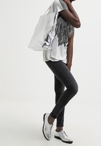 Silver tote bag made of metallic material, textured surface, carried alongside a grey graphic t-shirt and black skinny jeans, with silver shoes.