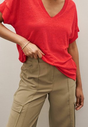 Persoon die een loszittend rood T-shirt met V-hals en een beige cargobroek met hoge taille draagt, met één hand die het shirt rechtzet en een armband om de pols.