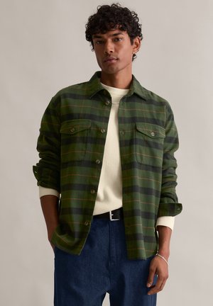 Zielona i czarna koszula w kratkę z dwiema kieszeniami na piersi, z zapięciem na guziki i kołnierzykiem, nałożona na jasny kremowy sweter.