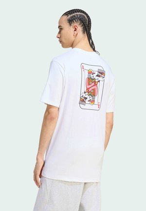 Junge Person mit geflochtenem Haar, die ein weißes T-Shirt mit einer bunten Spielkartengrafik mit einem König trägt, der Fast Food hält.