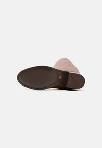 Semelle en caoutchouc marron avec une texture anti-dérapante, tige matelassée rose clair, design à bout pointu, détail de logo en relief près du talon.