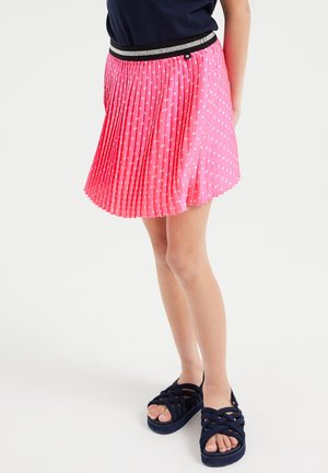 Fille portant une jupe plissée rose à pois blancs et des sandales bleu marine, debout sur un fond blanc uni.
