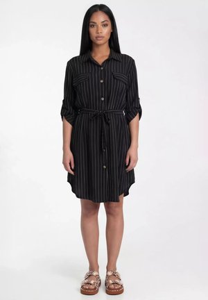 Kobieta stojąca, ubrana w czarną sukienkę koszulową w cienkie paski z podwiniętymi rękawami i wiązanym paskiem w talii, w połączeniu z beżowymi plecionymi sandałami na białym tle.