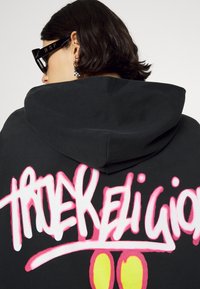 Persona con capelli corti scuri indossa una felpa con cappuccio nera con grande scritta in stile graffiti "True Religion" rosa e bianca sul retro e occhiali da sole neri.