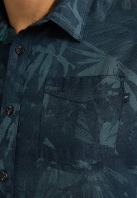 Camisa de manga corta de color azul oscuro con una tela texturizada y un diseño estampado de hojas. Incluye un bolsillo en el pecho y botones negros.