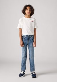 Camiseta de algodón blanca con logo rojo, jeans de mezclilla azules y zapatillas oscuras con suelas blancas; cabello rizado visible, de pie contra un fondo neutro.