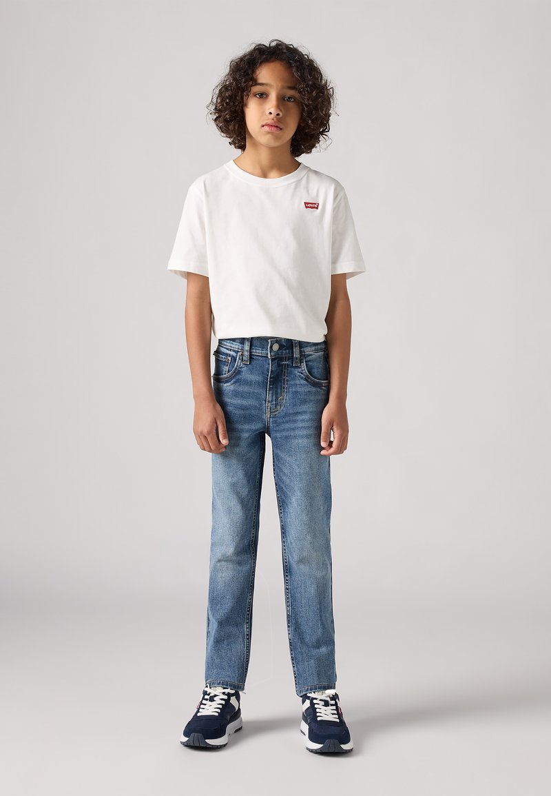 Camiseta de algodón blanca con logo rojo, jeans de mezclilla azules y zapatillas oscuras con suelas blancas; cabello rizado visible, de pie contra un fondo neutro.