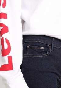 Sudadera blanca con texto rojo "Levi's" en la manga, combinada con jeans de mezclilla azul oscuro con costuras doradas y un pequeño bolsillo trasero.