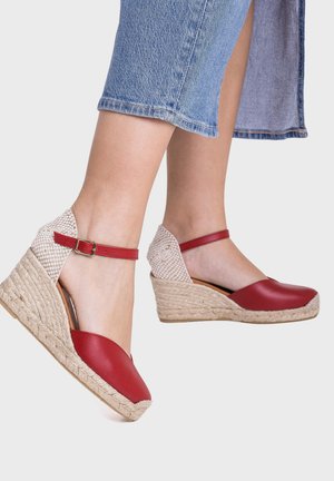 Pies con alpargatas cuña de cuero rojo con correas en el tobillo y tacones de yute tejido, combinados con vaqueros azules recortados.