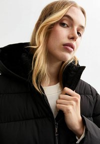 Doudoune noire avec un col haut, finition brillante, fermeture éclair et panneaux cousus. Portée par-dessus un pull de couleur claire.