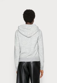 Persona con capelli ricci corti che indossa orecchini a cerchio dorati, una felpa con cappuccio grigia chiara e pantaloni di pelle neri, in piedi di spalle.