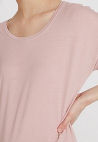 Kvinde iført en lys pink kortærmet skjorte med den ene hånd hvilende på hoften mod en ensfarvet baggrund.
