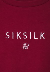 Bordová tkanina s bílým vyšívaným textem "SIKSILK" a malým dekorativním propleteným logem "SS" pod textem.
