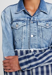 Osoba w dwukolorowej kurtce, z górną częścią z jasnoniebieskiego denimu i dolną z niebiesko-białej kratki, z założonymi na piersi skrzyżowanymi ramionami.