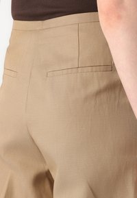 Pantaloni beige in tessuto liscio, con due tasche posteriori con cuciture orizzontali e vestibilità slim. Superficie testurizzata e colore neutro.