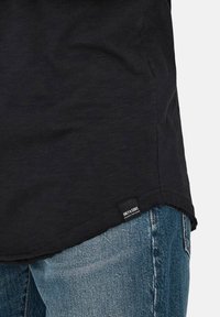 Zwart shirt met korte mouwen en een gebogen zoom, met een textuurafwerking, voorzien van een label aan de onderzijde links. Gemaakt in combinatie met vervaagde blauwe jeans.