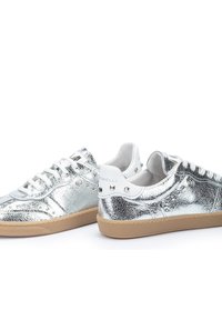 Zapatillas metálicas plateadas con un acabado texturizado, acentos blancos, cordones planos y una suela de goma. Presentan detalles con tachuelas en el talón.