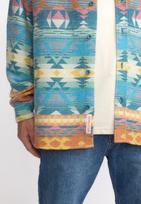 Meerkleurig geblokt overhemd met geometrische ontwerpen in blauw, roze en geel, voorzien van een label op de zoom. Katoenen textuur.