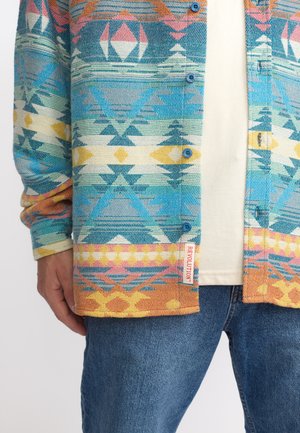 Meerkleurig geblokt overhemd met geometrische ontwerpen in blauw, roze en geel, voorzien van een label op de zoom. Katoenen textuur.
