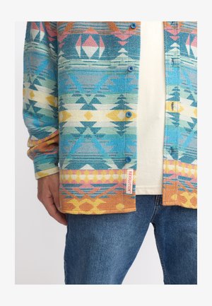 Multicolored patterned button-up shirt featuring geometric designs in blue, pink, and yellow, with a label on the hem. Cotton texture.