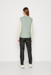 Jasnozielona pikowana kamizelka z wysokim kołnierzem, połączona z kremową koszulą z długimi rękawami i czarnymi skórzanymi spodniami. Model ma na sobie białe sneakersy.