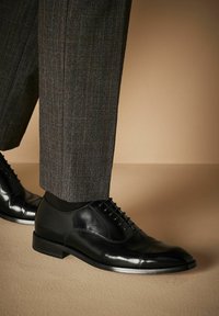 Zapatos de vestir de cuero negro con un acabado pulido, que presentan una puntera tipo cap toe, diseño con cordones y tacón bajo, combinados con pantalones grises a cuadros.