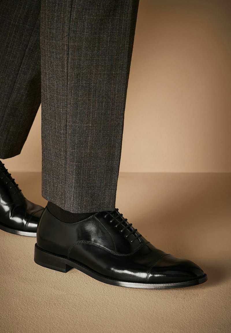 Zapatos de vestir de cuero negro con un acabado pulido, que presentan una puntera tipo cap toe, diseño con cordones y tacón bajo, combinados con pantalones grises a cuadros.