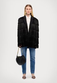 Zwarte oversized blazer met franjes en een gepolijste kraag, witte blouse, blauwe jeans en een zwarte gebogen handtas met een gouden schouderband.