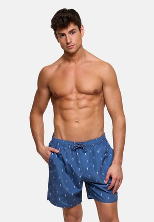 Hombre sin camiseta con cabello corto y castaño, vistiendo pantalones cortos de baño azules con pequeños patrones de caballitos de mar, de pie con una mano en el bolsillo contra un fondo blanco.