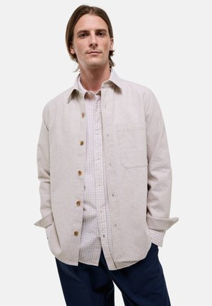 Jeune homme aux cheveux mi-longs portant une chemise beige boutonnée sur une chemise à carreaux et un pantalon bleu foncé, les mains dans les poches.