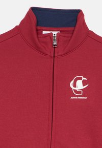 Κόκκινο μπουφάν με φερμουάρ και ψηλό γιακά, που δια θέτει εσωτερικό σε ναυτικό μπλε, λευκό λογότυπο στο στήθος και υφασμάτινη υφή για άνεση.