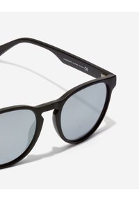 Black sunglasses with a rounded, cat-eye shape, grey tinted lenses, and matte texture. Frame features subtle metal accents on hinges.