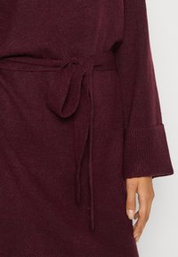 Burgunderfärgad stickad klänning med en knuten midjebälte, utrustad med lösa ärmar och en ribbad textur vid ärmsluten. Mjukt, varmt tyg.