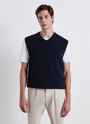 Uomo che indossa un maglione smanicato blu navy con scollo a V sopra una camicia bianca a maniche corte e pantaloni beige, in piedi davanti a uno sfondo semplice.