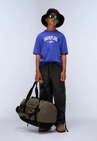 Αγόρι που φοράει μαύρο καπέλο μπέιζμπολ και γυαλιά ηλίου, μπλε t-shirt Napapijri, μαύρο cargo παντελόνι, κρατώντας μια μεγάλη ελαιοπράσινη τσάντα duffel με το λογότυπο της νορβηγικής σημαίας.