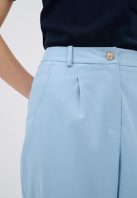 Hellblaue Hosen mit einer glatten Textur, einem einzelnen Bundfalten, seitlichen Taschen und einem goldenen Knopf am Bund.